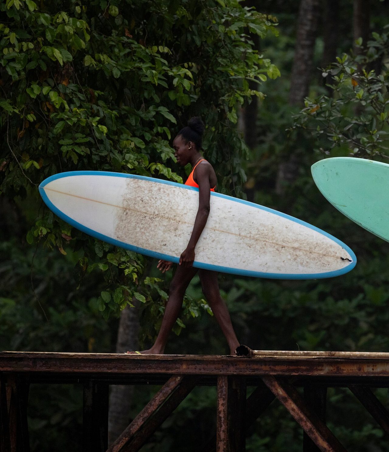 Mujer en São Tomé caminando sobre un puente de hierro, con una tabla de surf en la mano, con vegetación detrás
