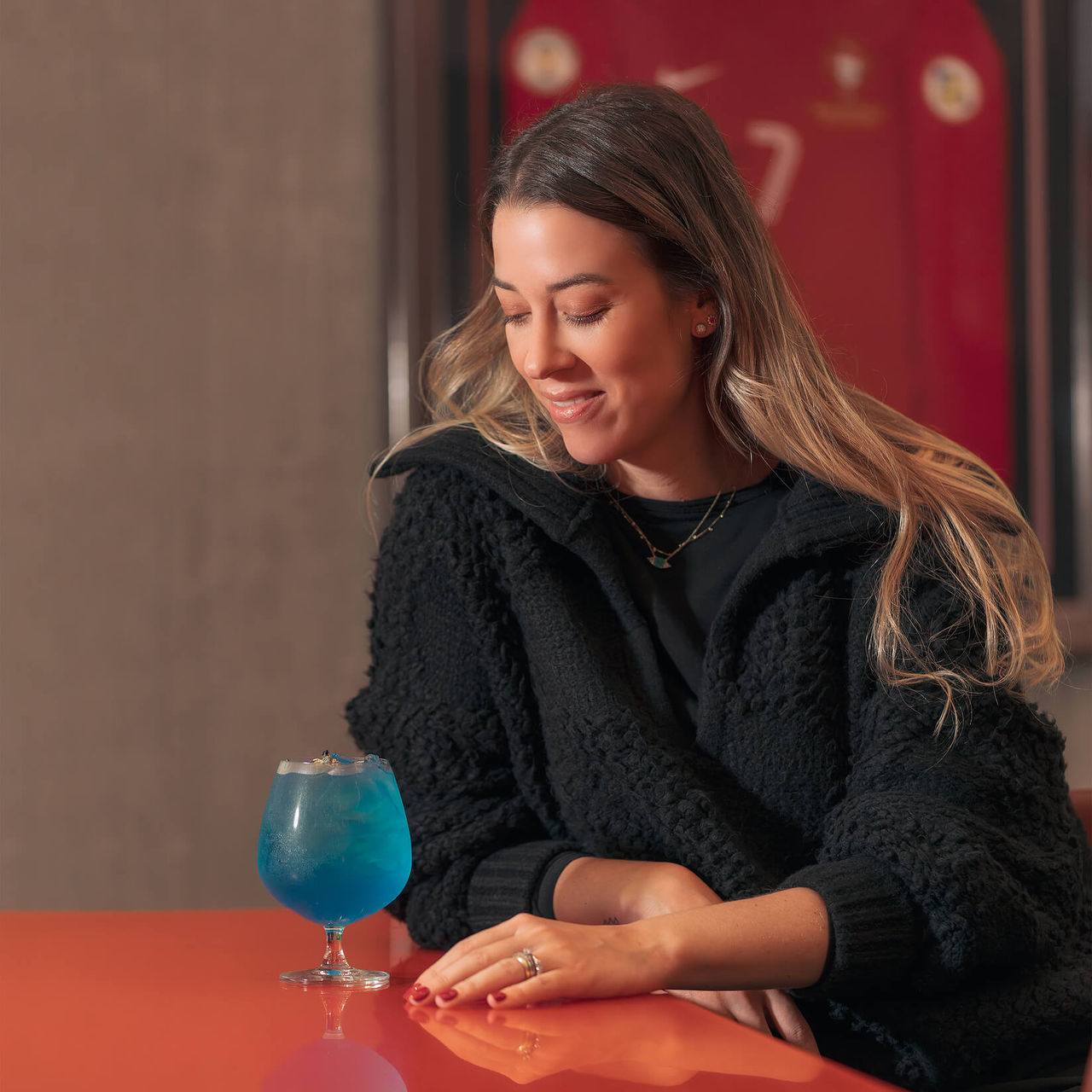Chica bebiendo un cóctel, sentada en una mesa, con la camiseta de Cristiano Ronaldo colgada en la pared