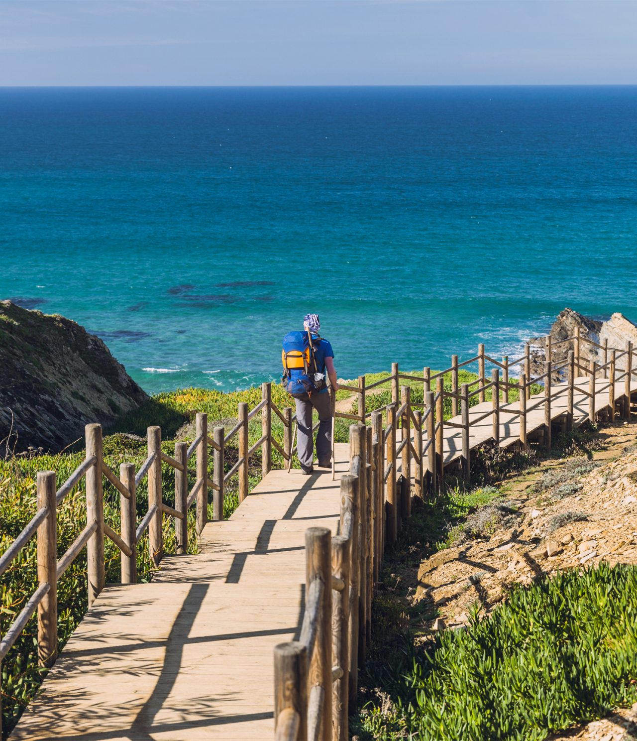Hombre caminando por un pasadizo de madera, hacia una playa en Alentejo, con aguas cristalinas