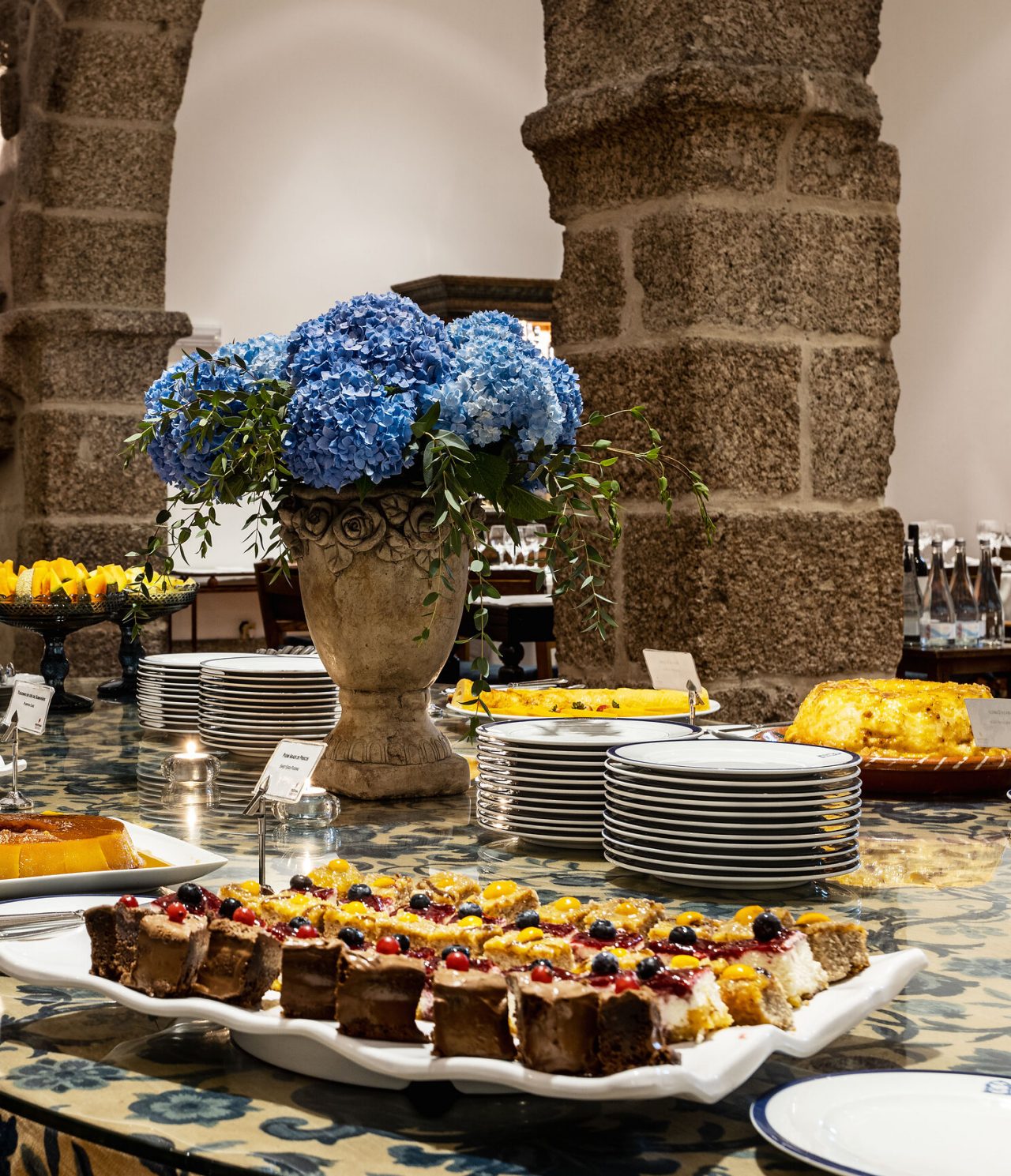 Mesa decorada con una variedad de dulces, desde pasteles y tartas hasta frutas frescas, en una sala con muebles antiguos