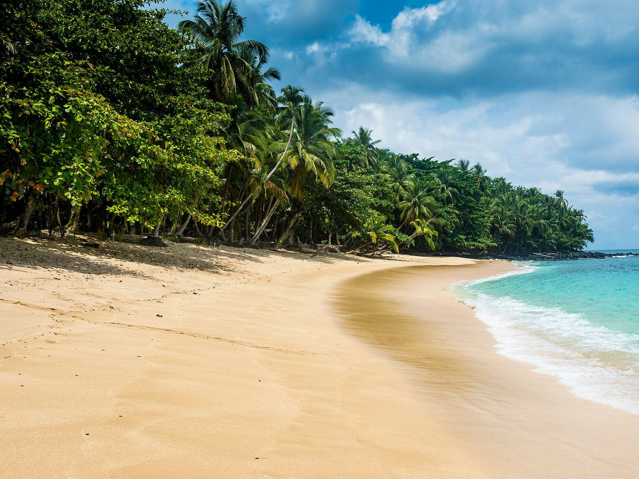 Praia deserta em São Tomé, com mar muito azul e calmo, e uma densa vegetação a rodear a praia