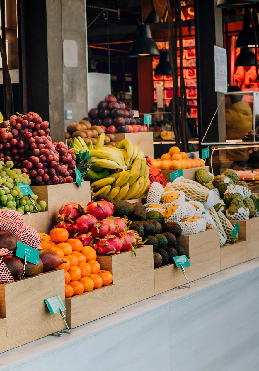Banca com frutas variadas no Mercado de San Miguel, balança e candeeiros de teto pretos, com um balcão de vidro