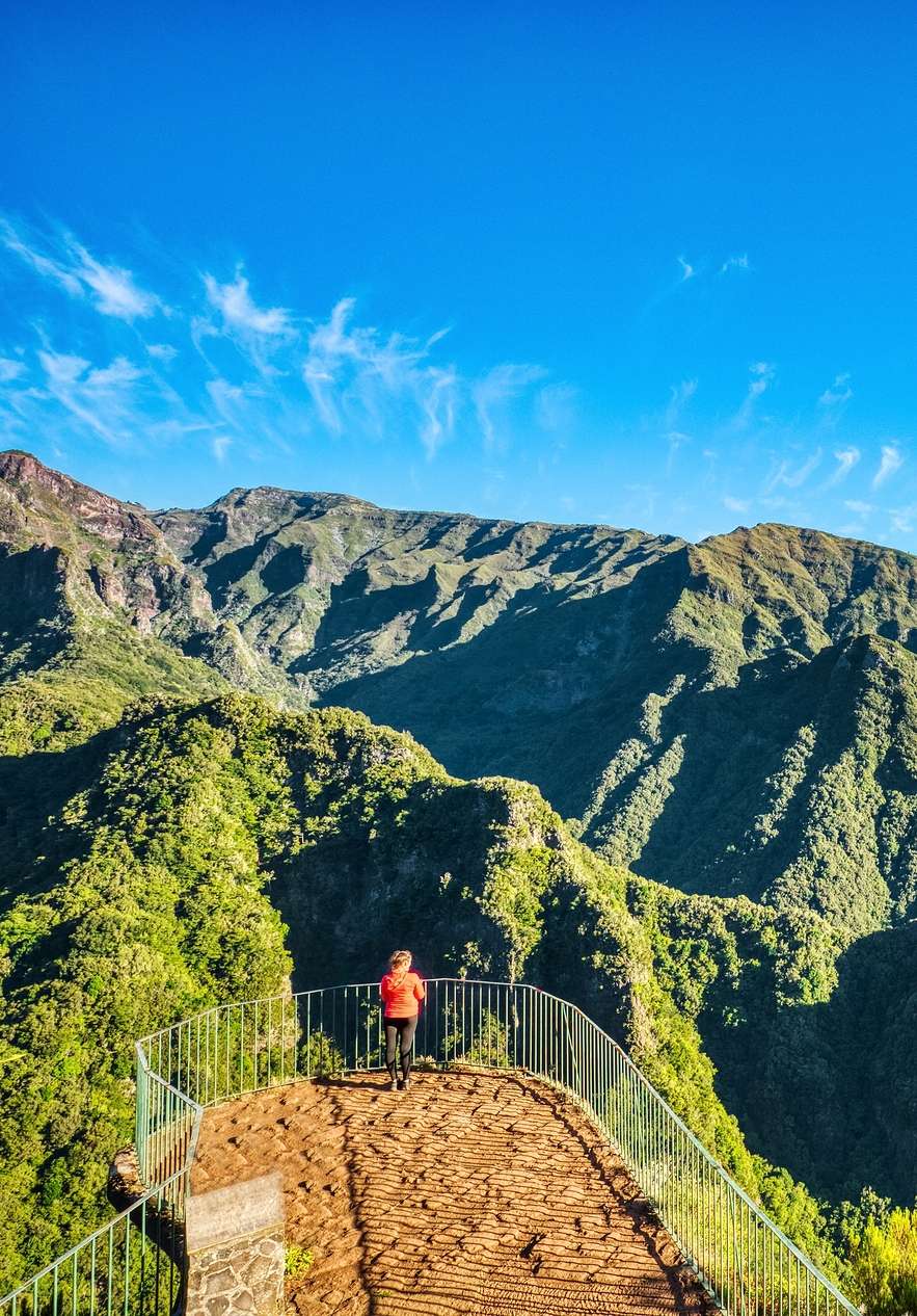 Pessoa a caminhar, que parou para apreciar a paisagem montanhosa verdejante, num miradouro na Madeira