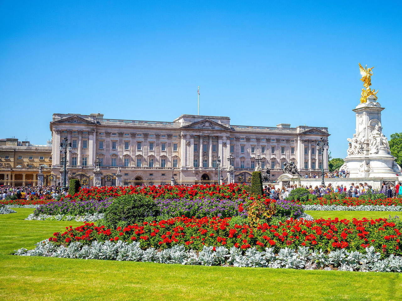 A imponente fachada do Palácio de Buckingham em Londres, com uma estátua dourada e um jardim florido em frente