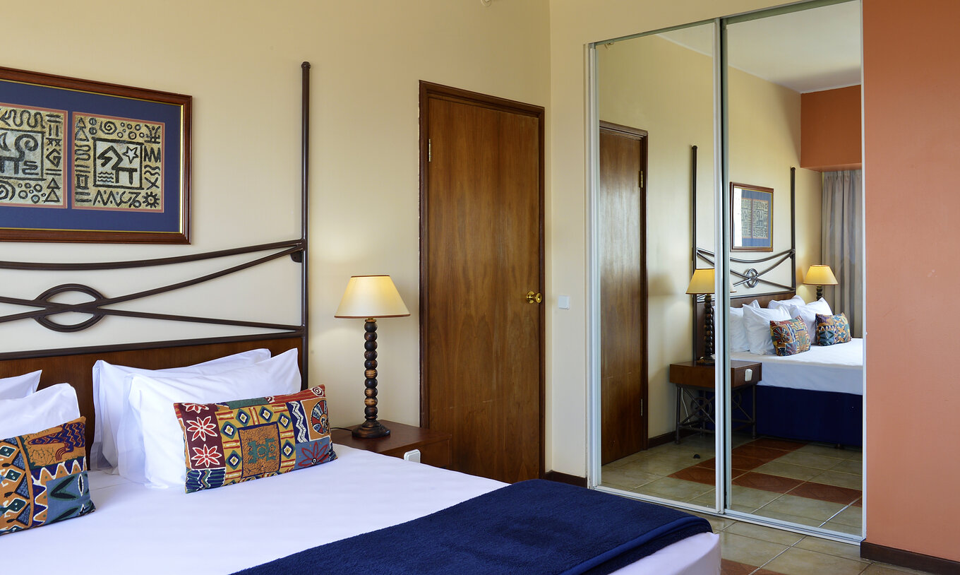 The Standard Double of Pestana Rovuma has a double bed, an African-themed painting, and a wardrobe with a large mirror