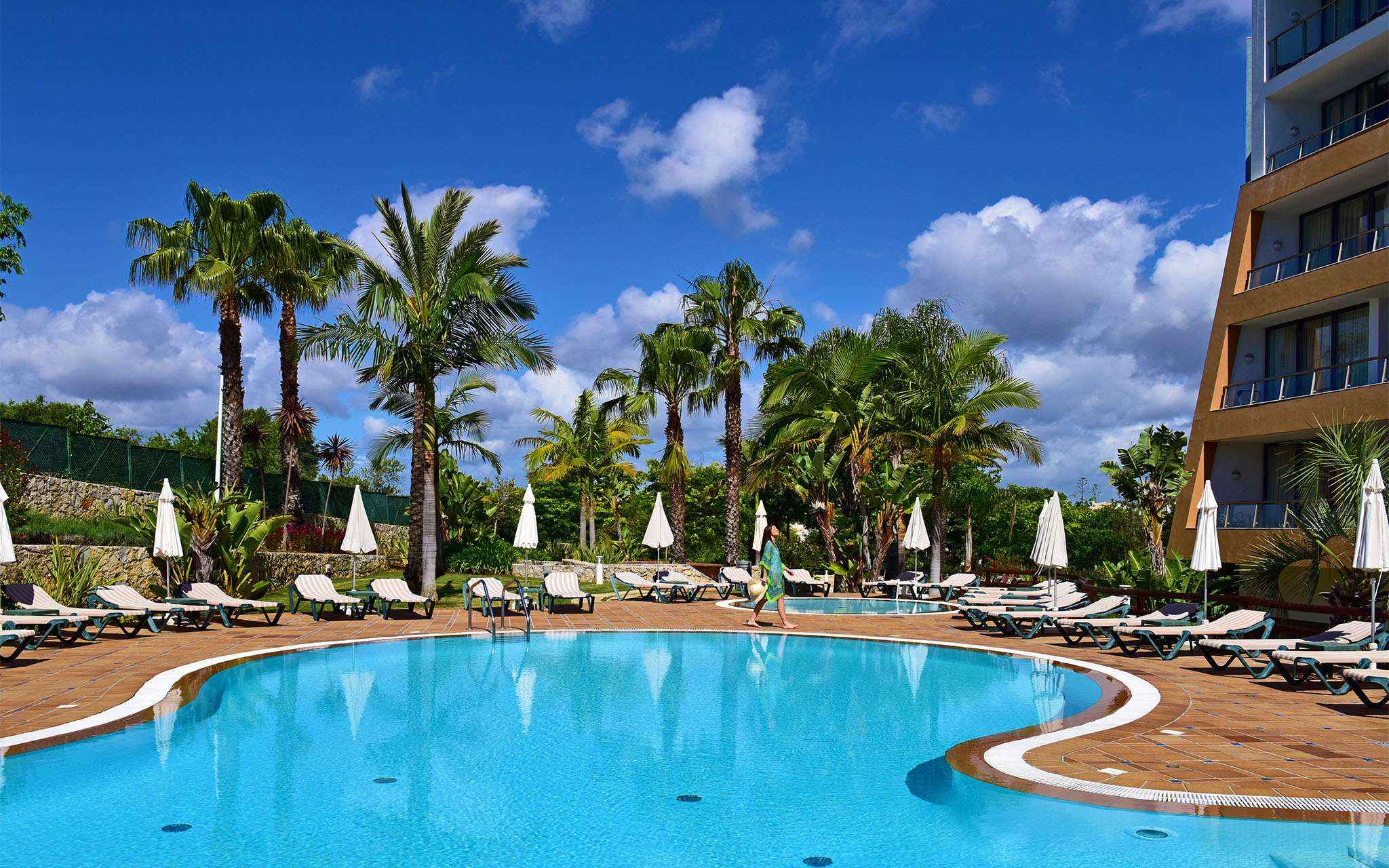 Outdoor pools with sun loungers, umbrellas, and palm trees, with a lady walking and the building on the side