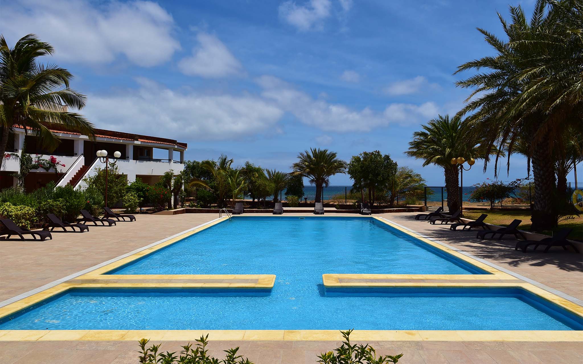 Vista de la piscina exterior de Pestana Trópico, con palmeras y tumbonas alrededor, y el mar al fondo