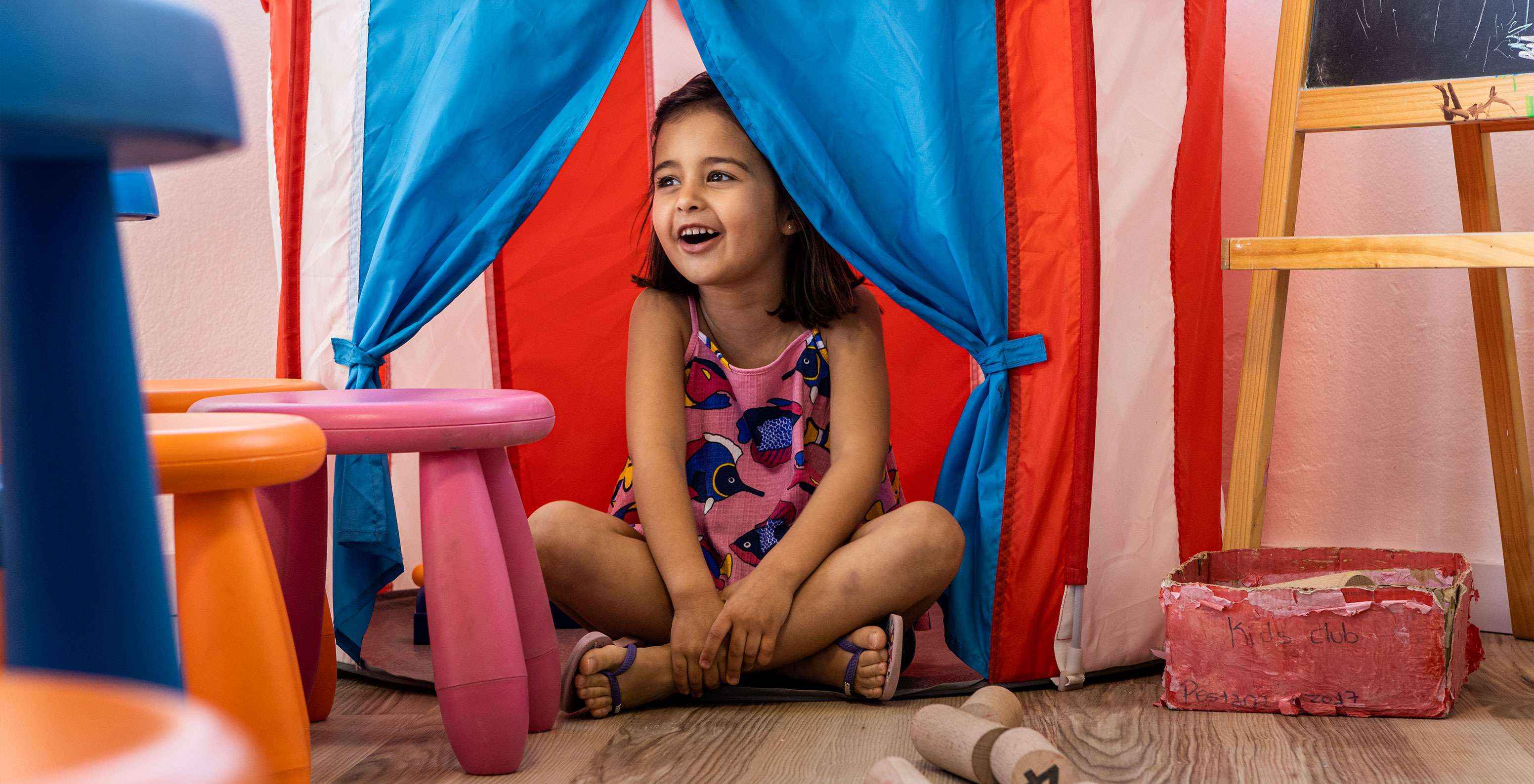 Un niño juega sonriente en el Kids Club, escondiéndose en una tienda roja y azul