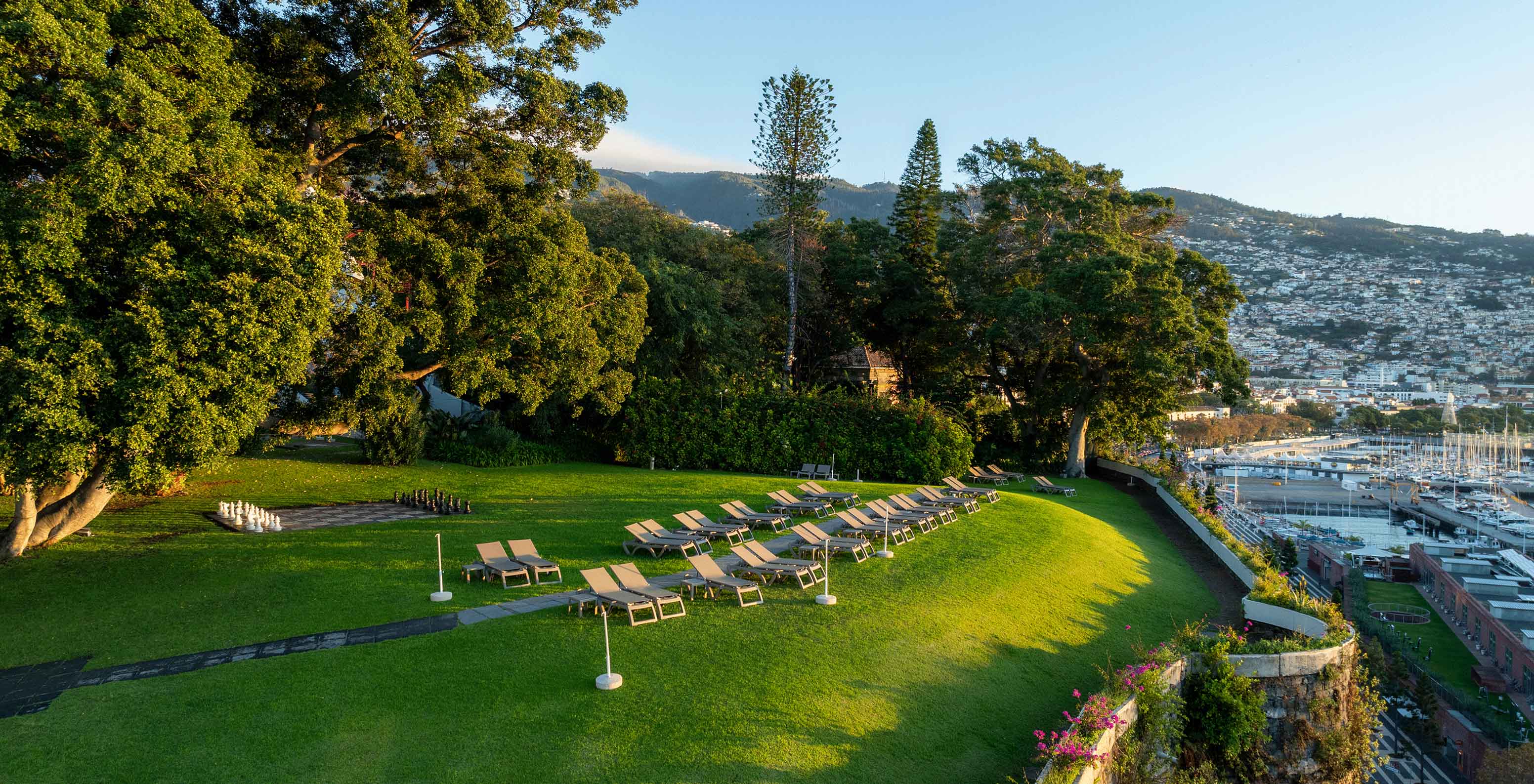 Vista del jardín del Pestana Casino Park al atardecer, con una ciudad al fondo y un crucero anclado en el mar.