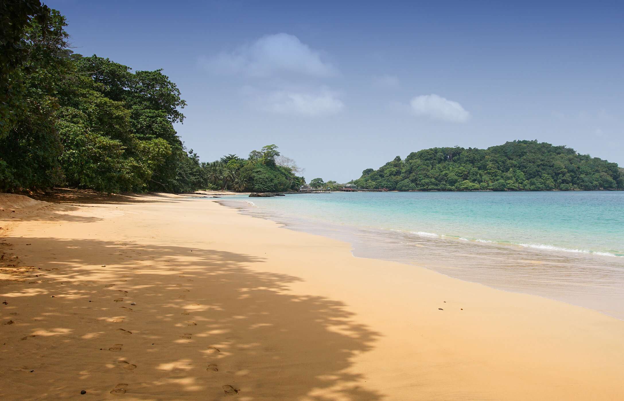 Vista de la playa de Coco en Santo Tomé y Príncipe, con arena dorada, aguas tranquilas cristalinas y vegetación alrededor.