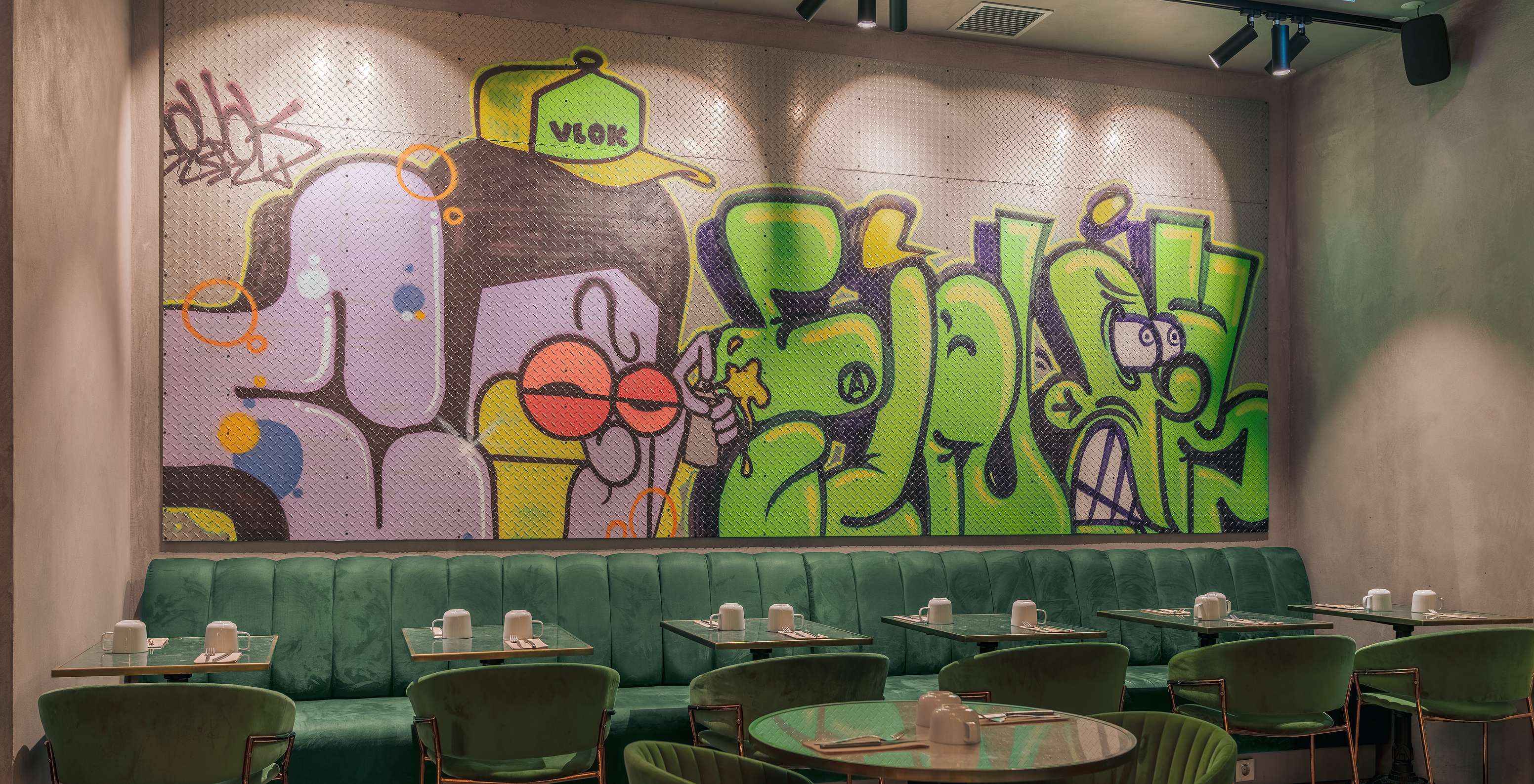 Restaurant met groene tafels, stoelen en banken, met witte borden en een gekleurde muur met graffiti