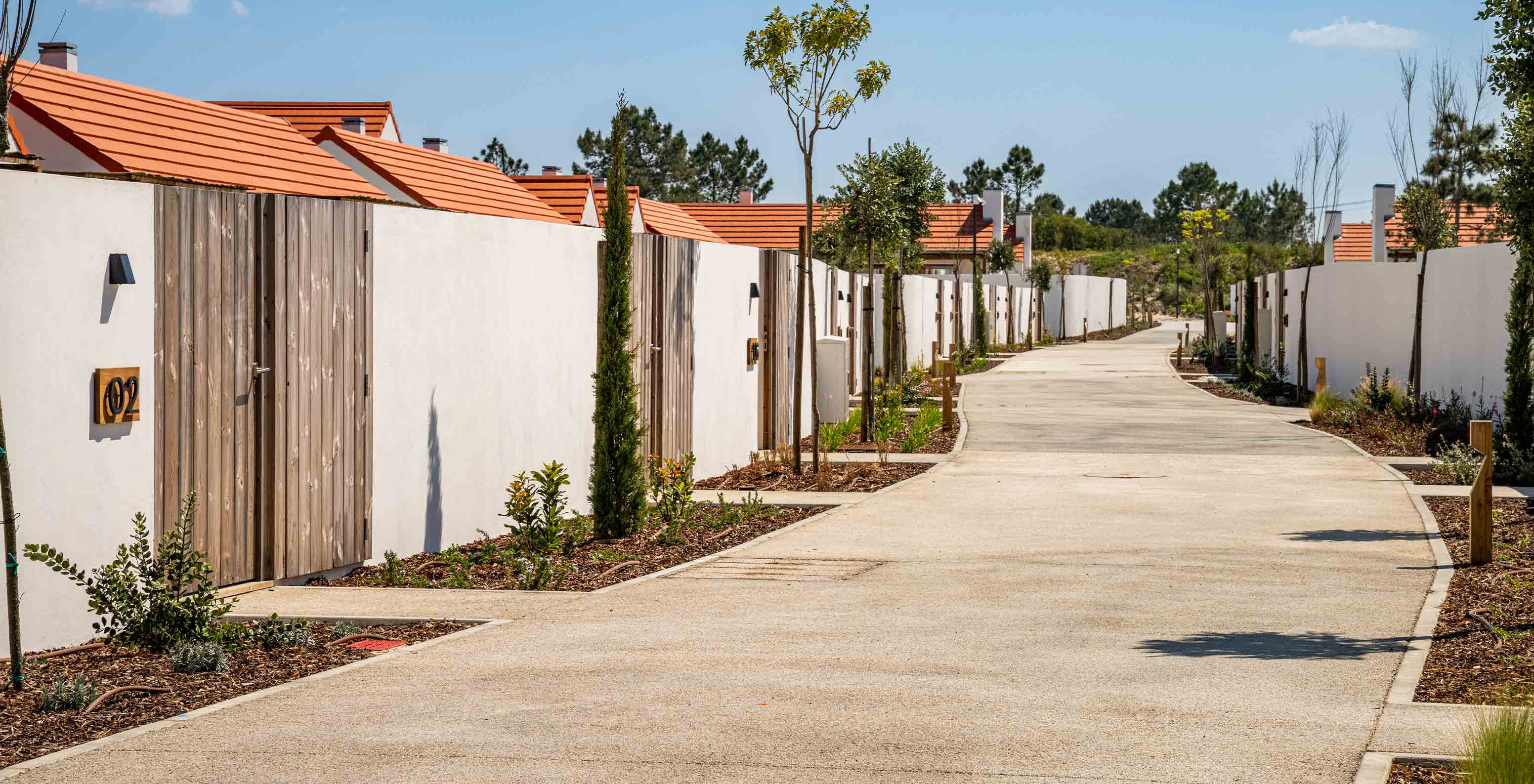 Rua pavimentada, ladeada por plantas, muros brancos e portões de madeira, entre villas com vegetação no fundo