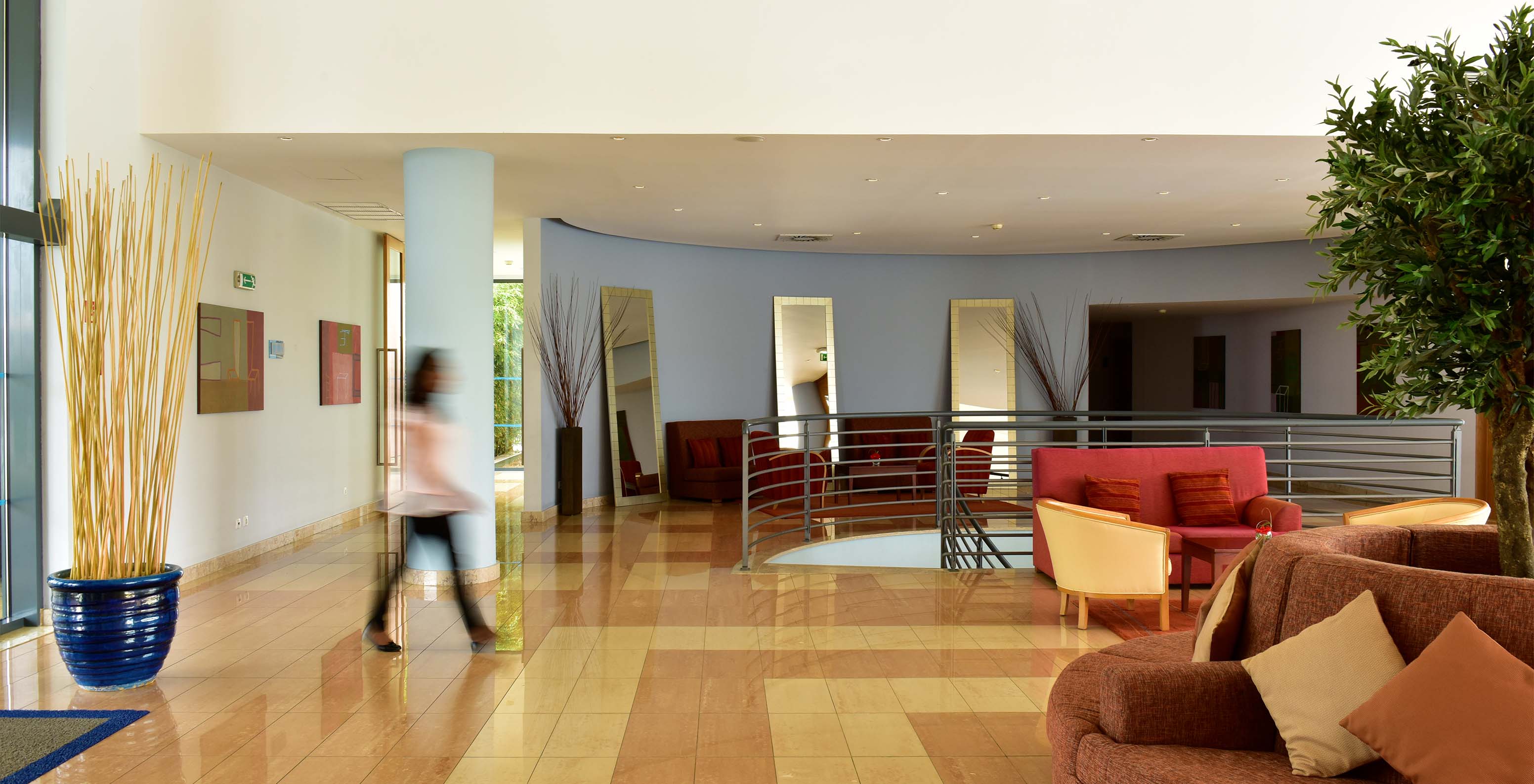 Entrada do hotel com sofás vermelhos e laranjas, com escadas metálicas e uma senhora a andar