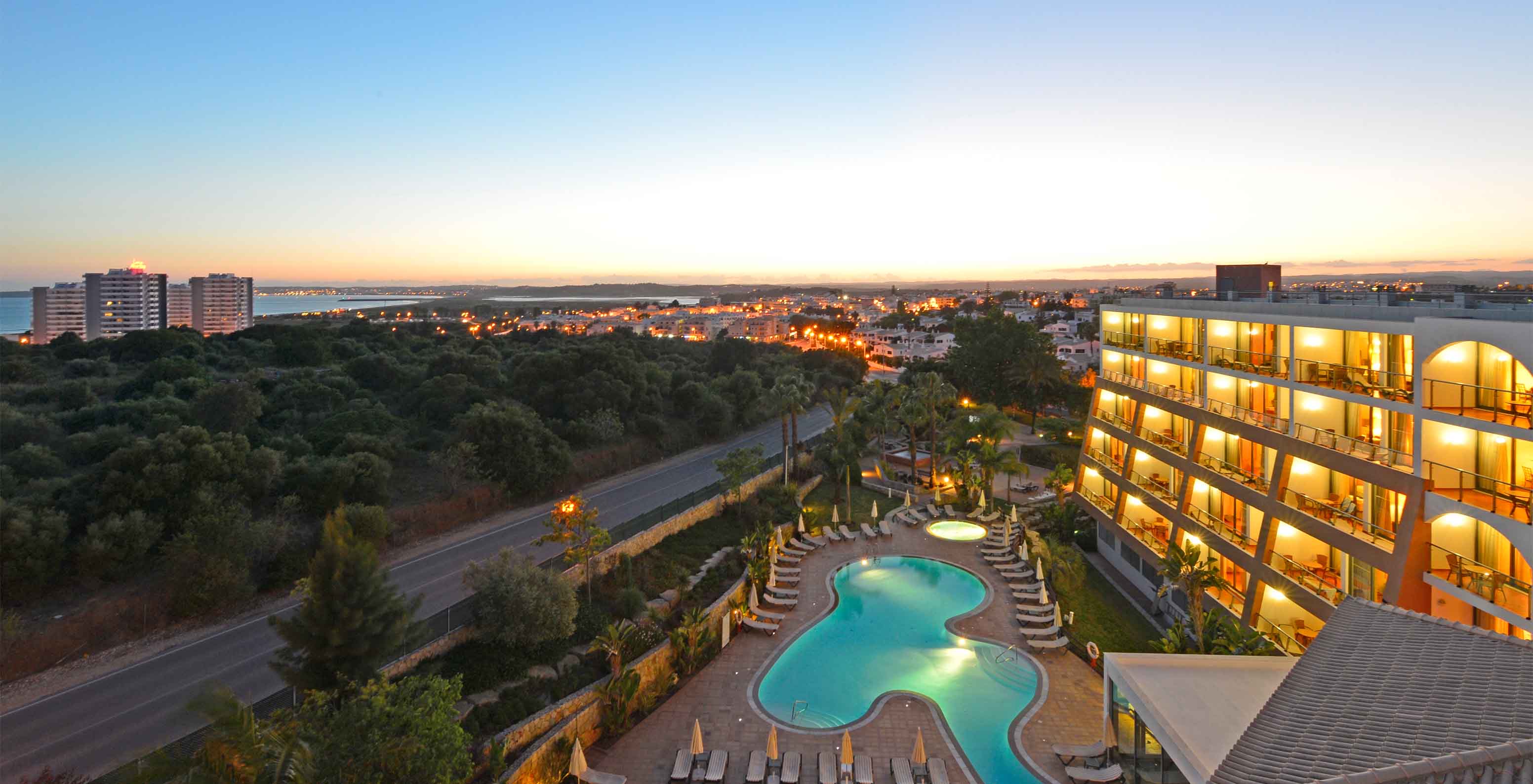 Vista de cima do Pestana Alvor Park, com o edifício e a piscina no pôr do sol, com o Alvor no fundo