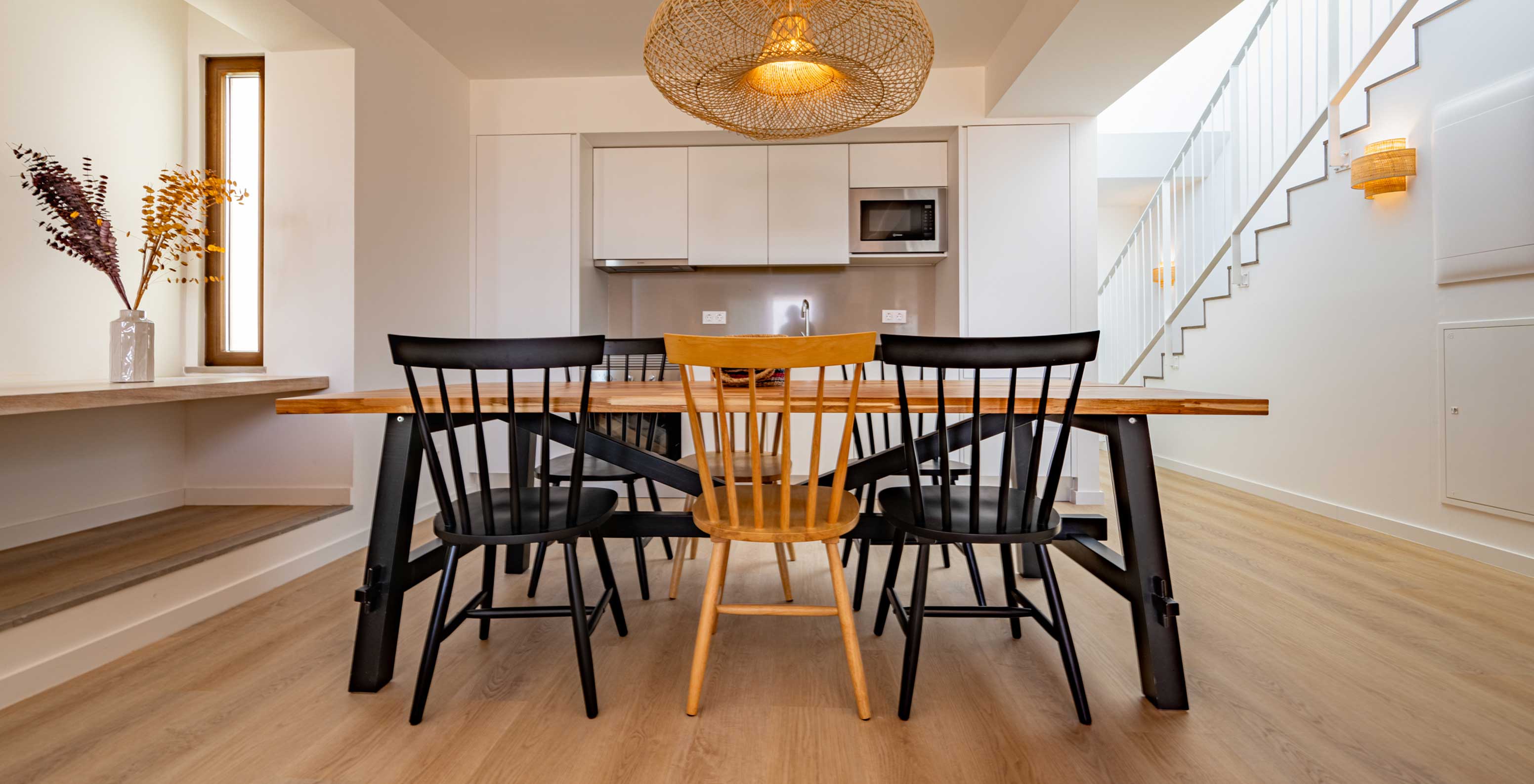 Mesa de jantar em madeira com quatro cadeiras, cozinha moderna ao fundo e escada branca ao lado