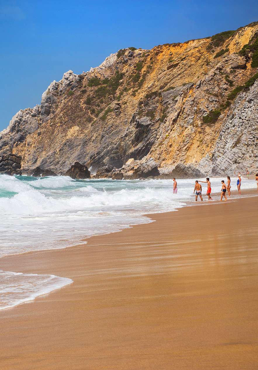 Strand in Sintra, met golven die breken tegen de kust, mensen die het water in gaan en rotsen op de achtergrond