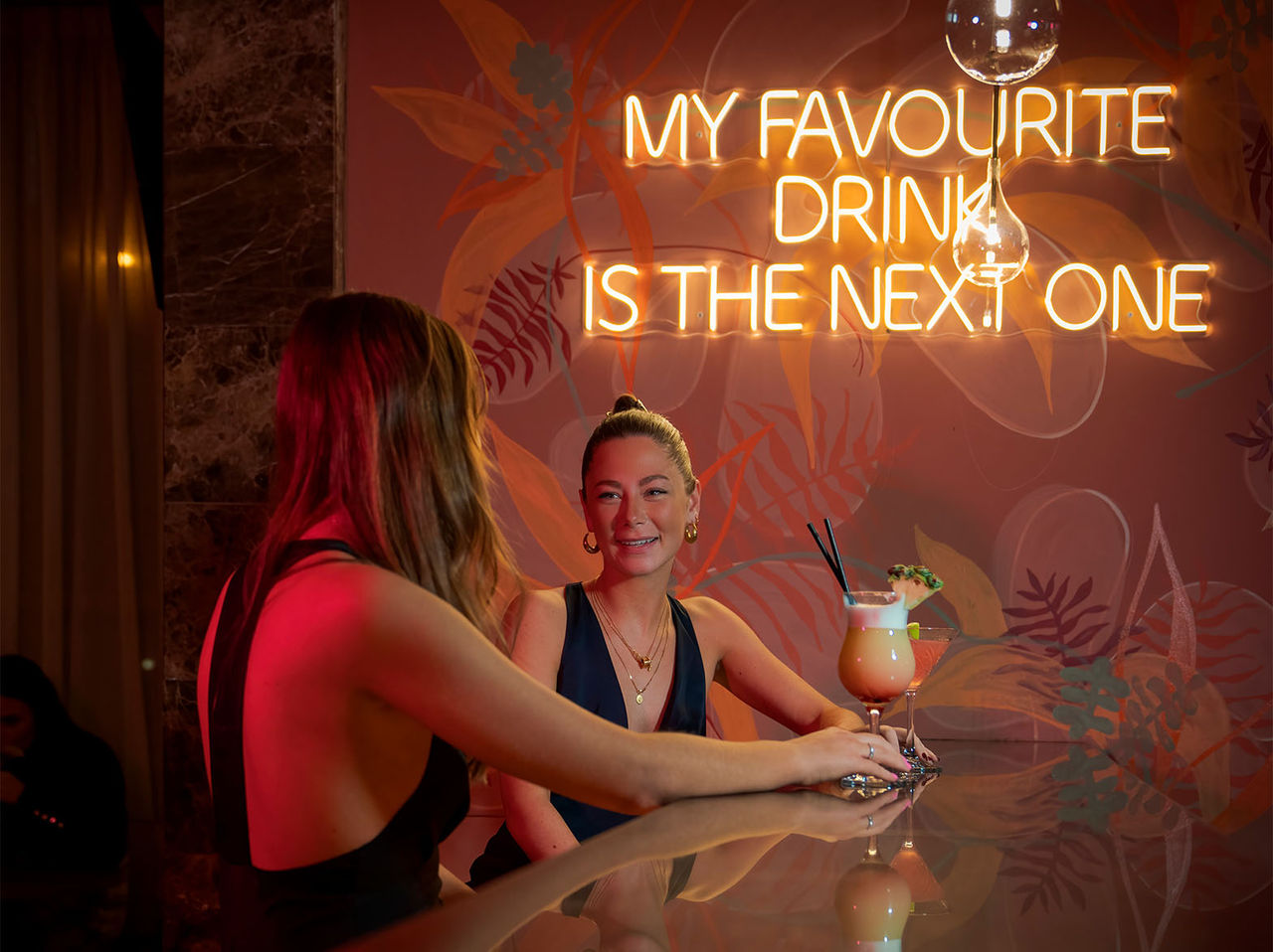 De bar The Bridge, van het 4-sterrenhotel in Londen, heeft twee vrouwen aan de bar met cocktails en een neonzin op de muur
