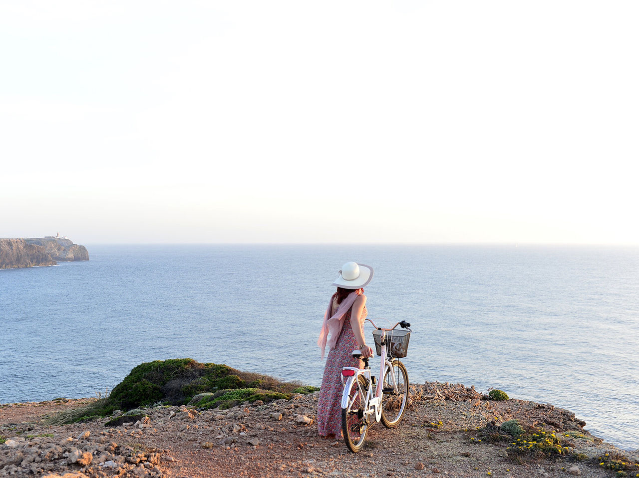 Young woman with a hat and a bicycle in hand, enjoying the view of the sea from a cliff in the Algarve