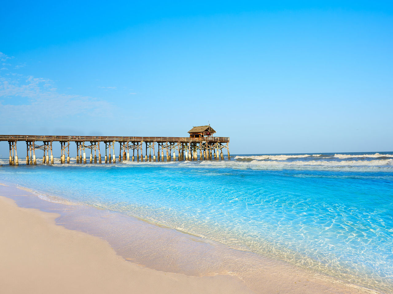 Long wooden pier over crystal-clear water, offering a view of a white sandy beach with a relaxing sea