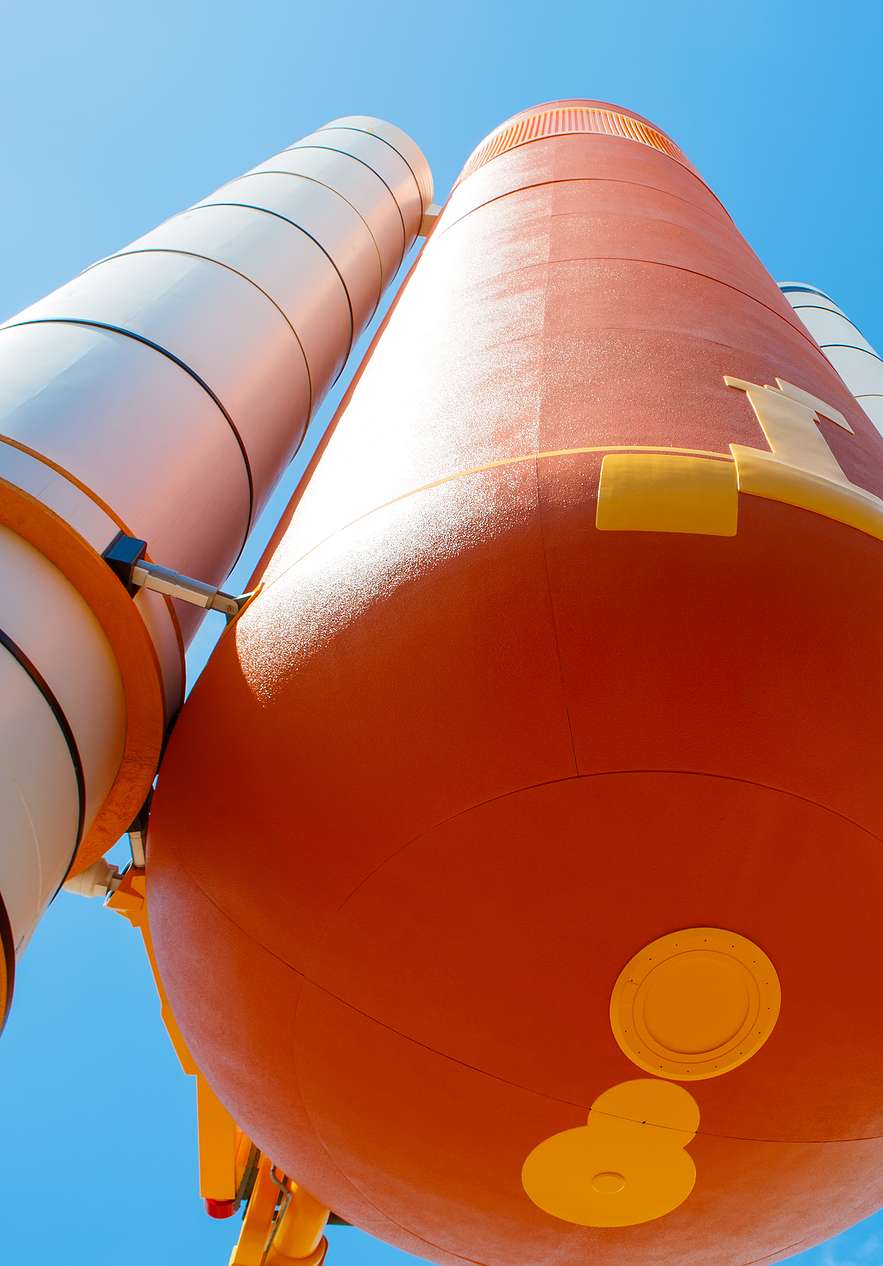 View from below of an external tank of an orange and white rocket with a blue sky and some clouds in the background