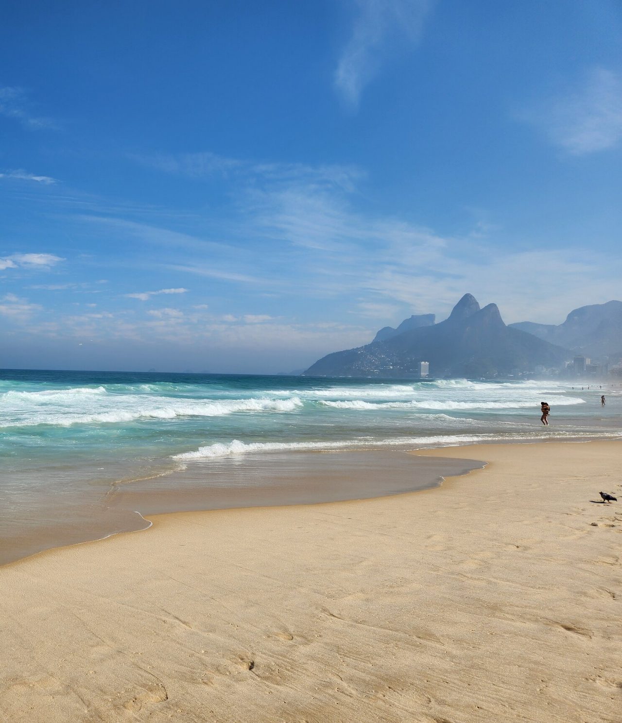 View of a beach in Rio de Janeiro, with white sand, turquoise water, mountains, and people by the water