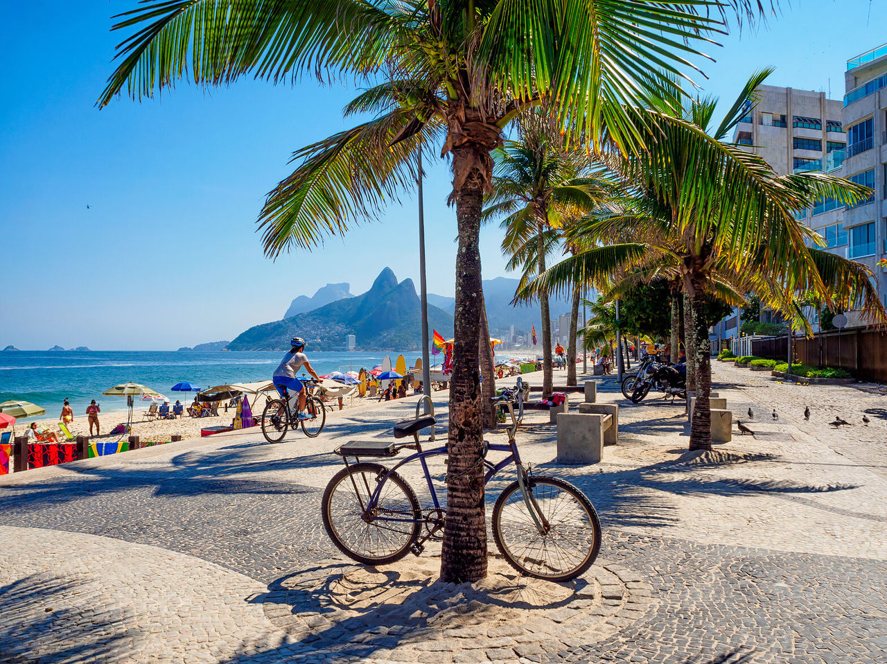 View of Ipanema beach, with people sunbathing, cyclists, and the Dois Irmãos hill in the background